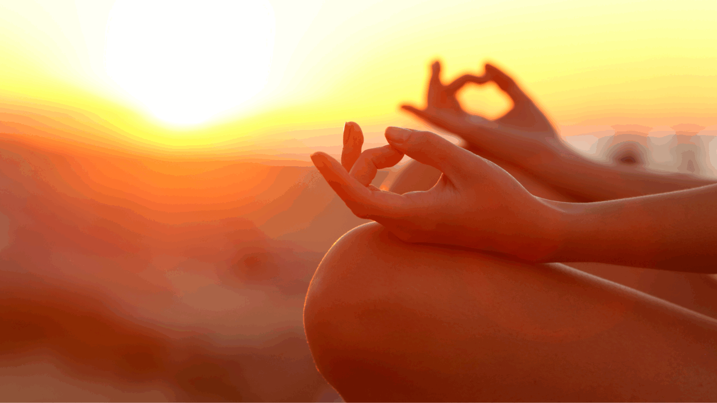 Feminine hands in a meditative pose with an orange sunset in the background.