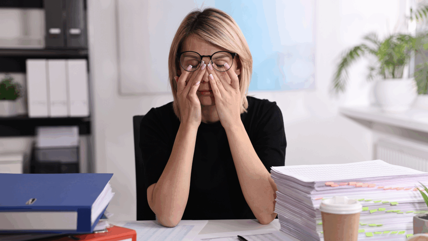 Woman sat at a desk with piling paperwork with her head in her hands.