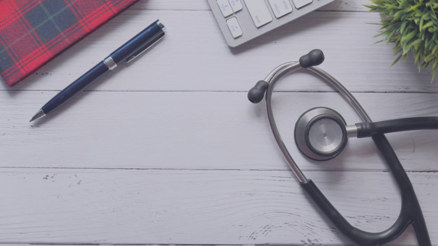 Birds eye view of a clinic desk with notepad, pen, laptop, plant and stethoscope in view.